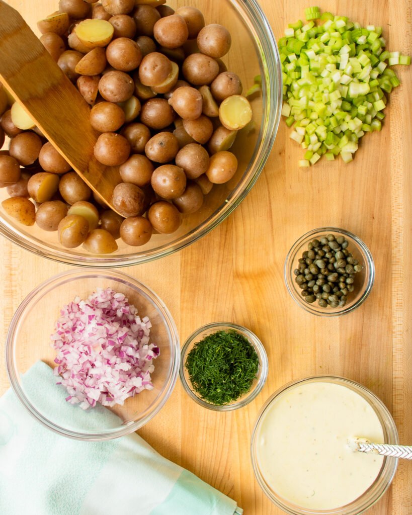 Looking down at a bowl of baby red potatoes, diced onion, celery, capers, dill and dressing.