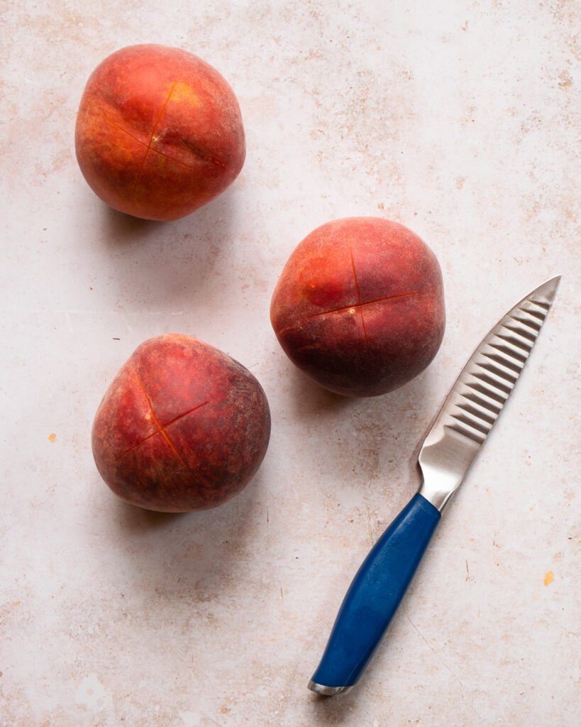 Three peaches with scored ends on a counter with a blue handled paring knife.