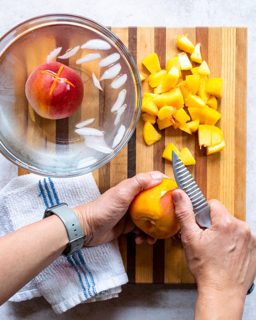 Two hands peeling a peach with another peach floating in an ice bath and diced peaches on a cutting board.