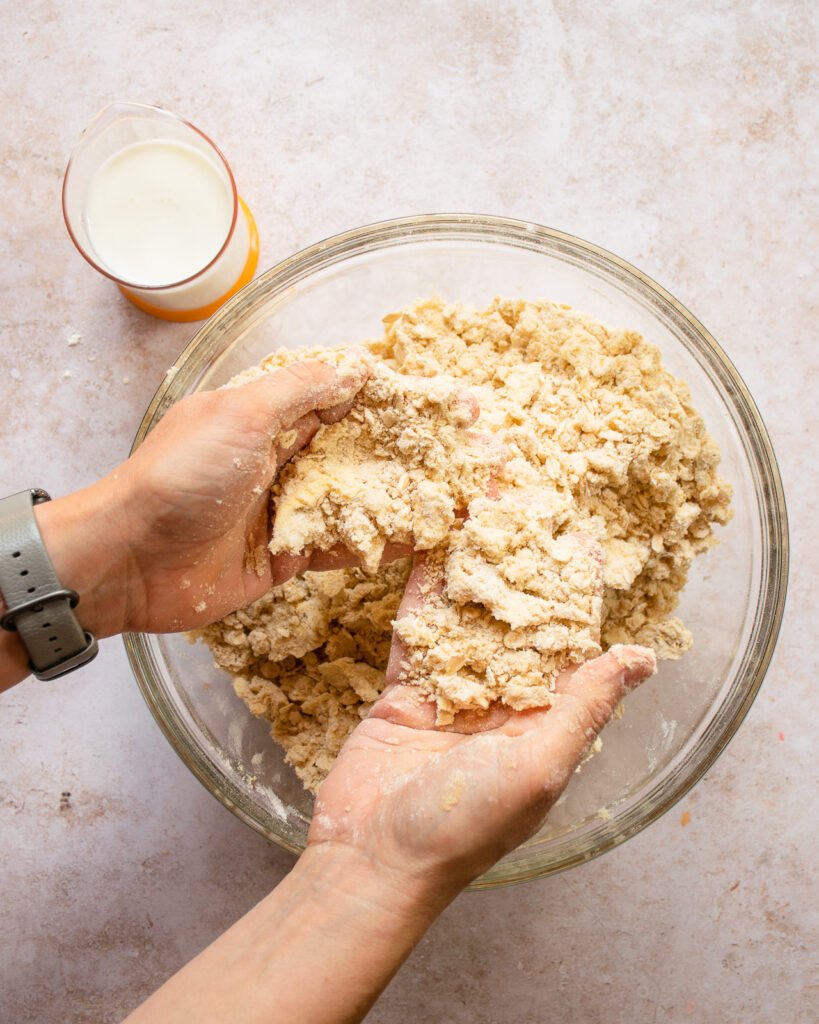 Hands mixing butter into dry ingredients with a measuring glass of butter milk next to it.