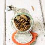 Pickling Spice in a glass jar with the lid off on a white table.