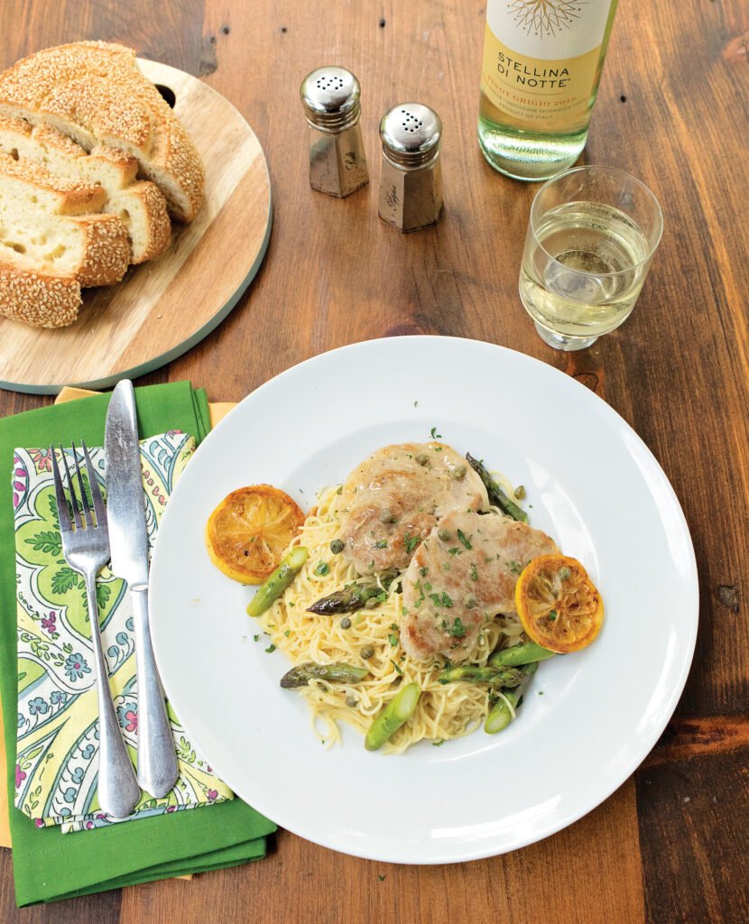 Pork Picatta on a bed of pasta with asparagus spears on a wooden table with bread, salt and pepper and white wine.