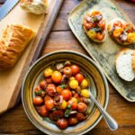 Roasted cherry tomatoes in a brown bowl with sliced baguette and crostini near by.