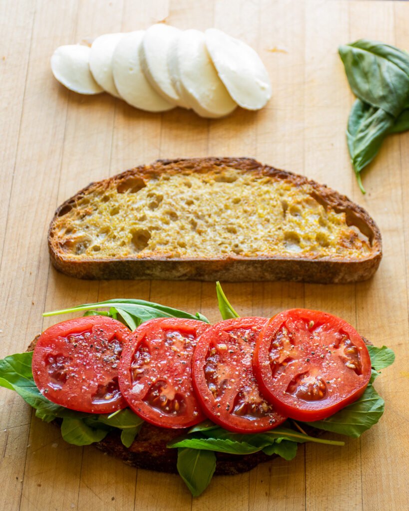 A sandwich being built on a wooden cutting board with tomato slices on a piece of bread and other ingredients around.