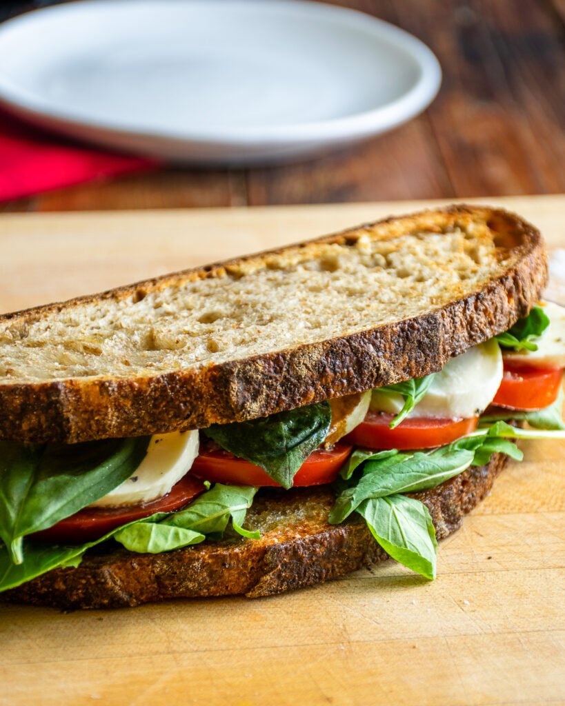 Caprese sandwich on a wooden cutting board with a white plate and red napkin in the background.