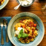 A blue bowl of cavatelli pasta with roasted cherry tomatoes, pancetta and burrata on a wooden table.