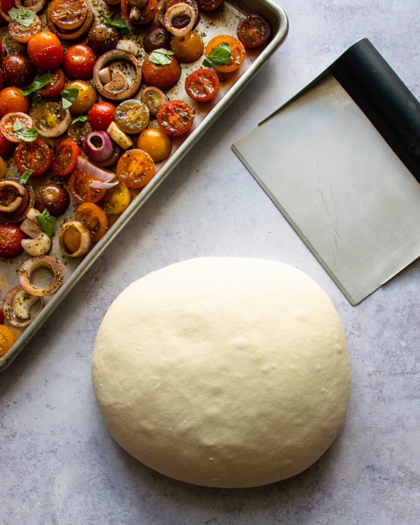 Focaccia dough ball on a counter with a bench scraper and a baking sheet of cherry tomatoes.