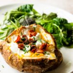 A baked potato with sour cream, chives and bacon on a white plate with salad greens in the background.