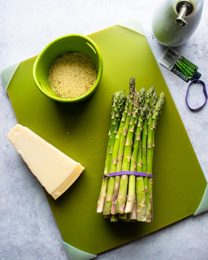 A bunch of asparagus, a bowl of breadcrumbs and a block of Parmesan cheese on a green cutting board.