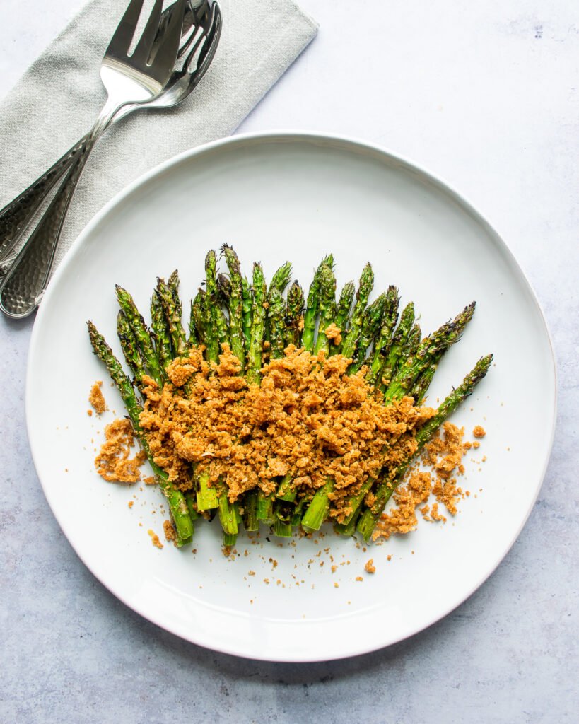Asparagus with crispy parmesan breadcrumbs on a white plate with serving utensils next to it.