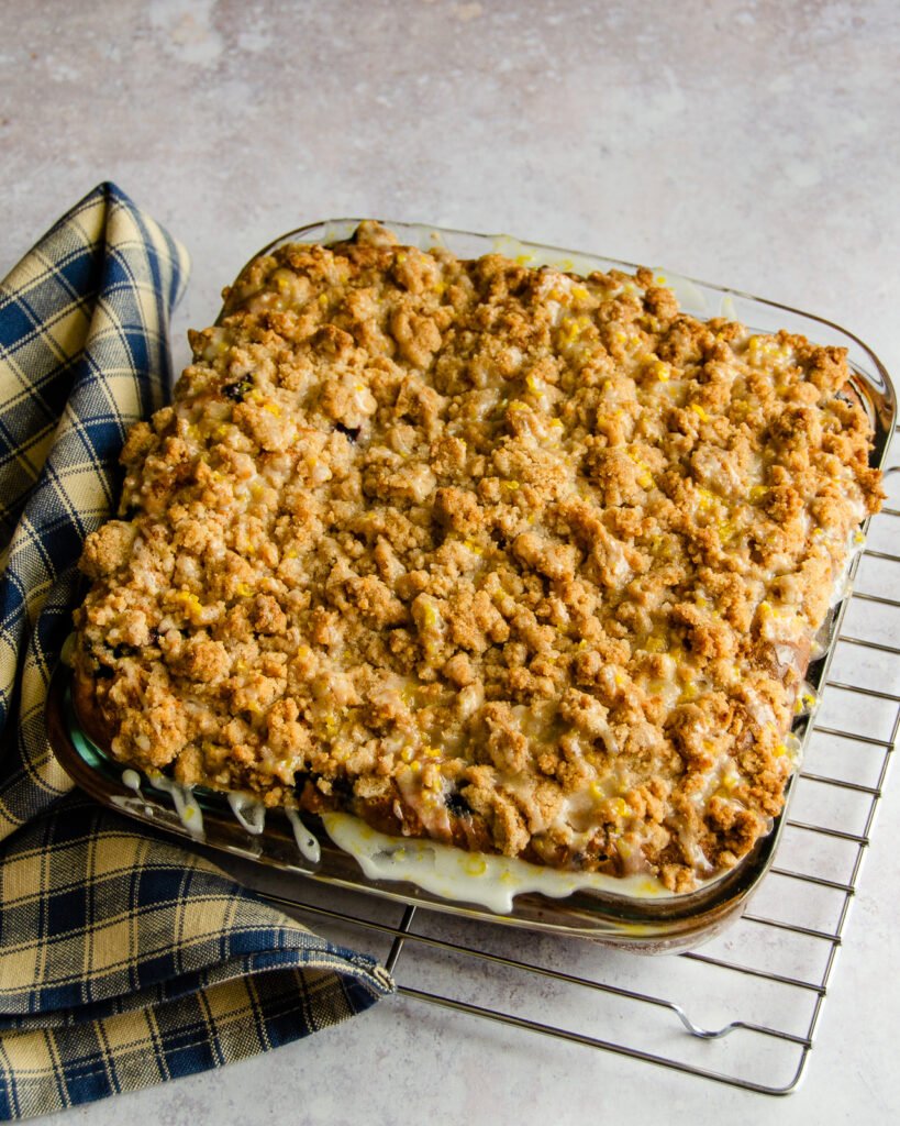 Blueberry crumb coffee cake in a glass baking dish on a wire rack with a blue checkered towel near by.
