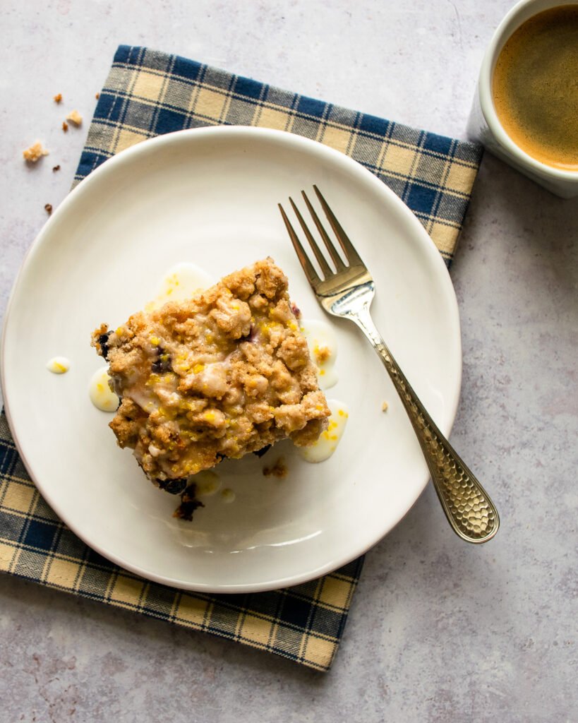 Looking straight down on a piece of blueberry coffee cake on a white plate with a fork and a cup of coffee.