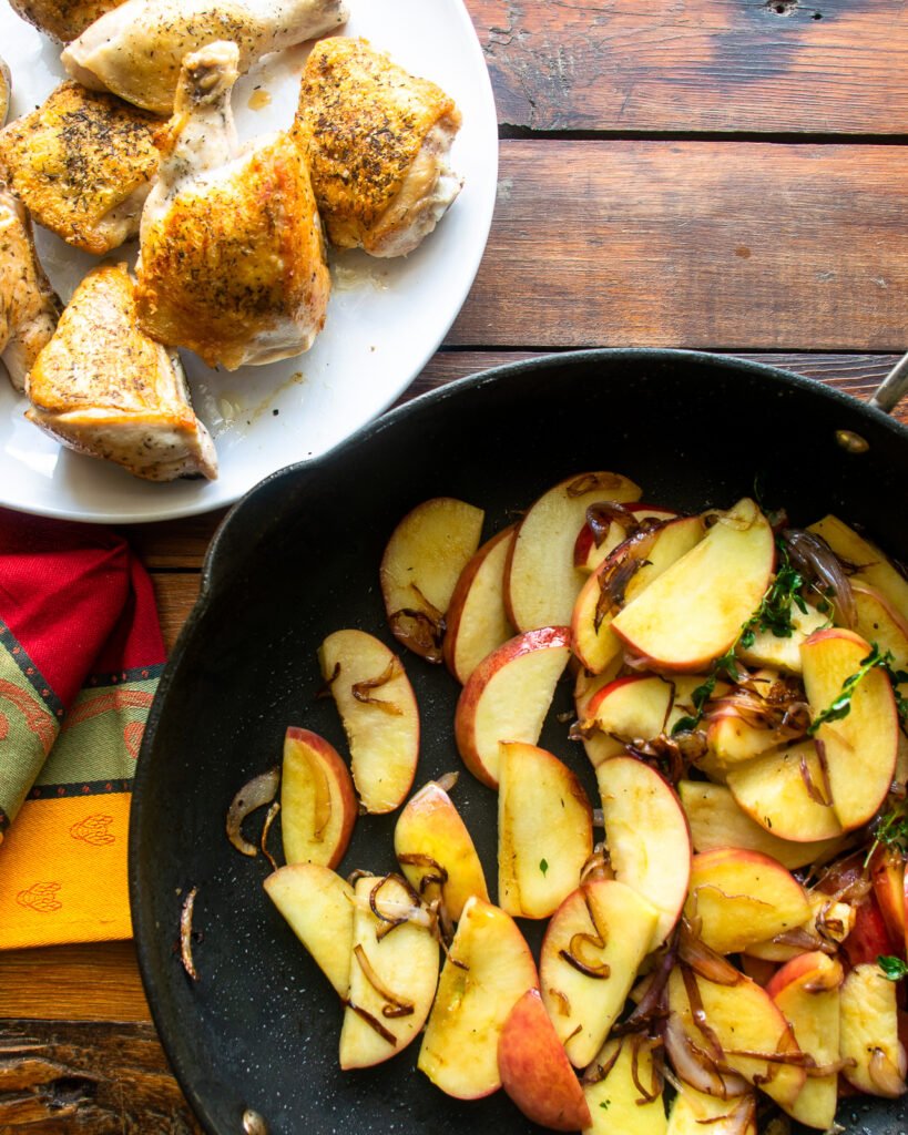 Apples, onions and thyme cooking in a pan with a plate of browned chicken next to it.