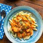 A light blue ceramic bowl with penne and salmon with tomato vodka cream sauce on a wooden table with a blue napkin.