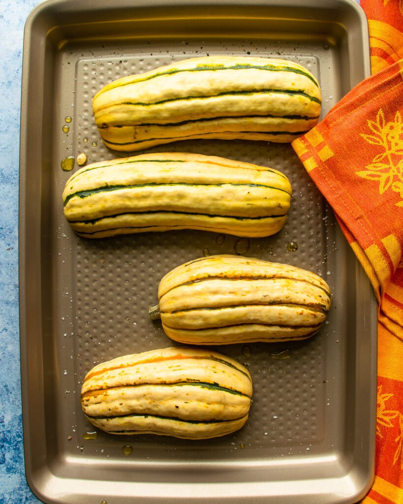 Four delicata squash halves cut side down on a baking sheet with an orange towel.
