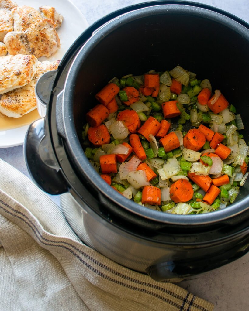 Vegetables cooking in a pressure cooker with a plate of browned chicken next to it.