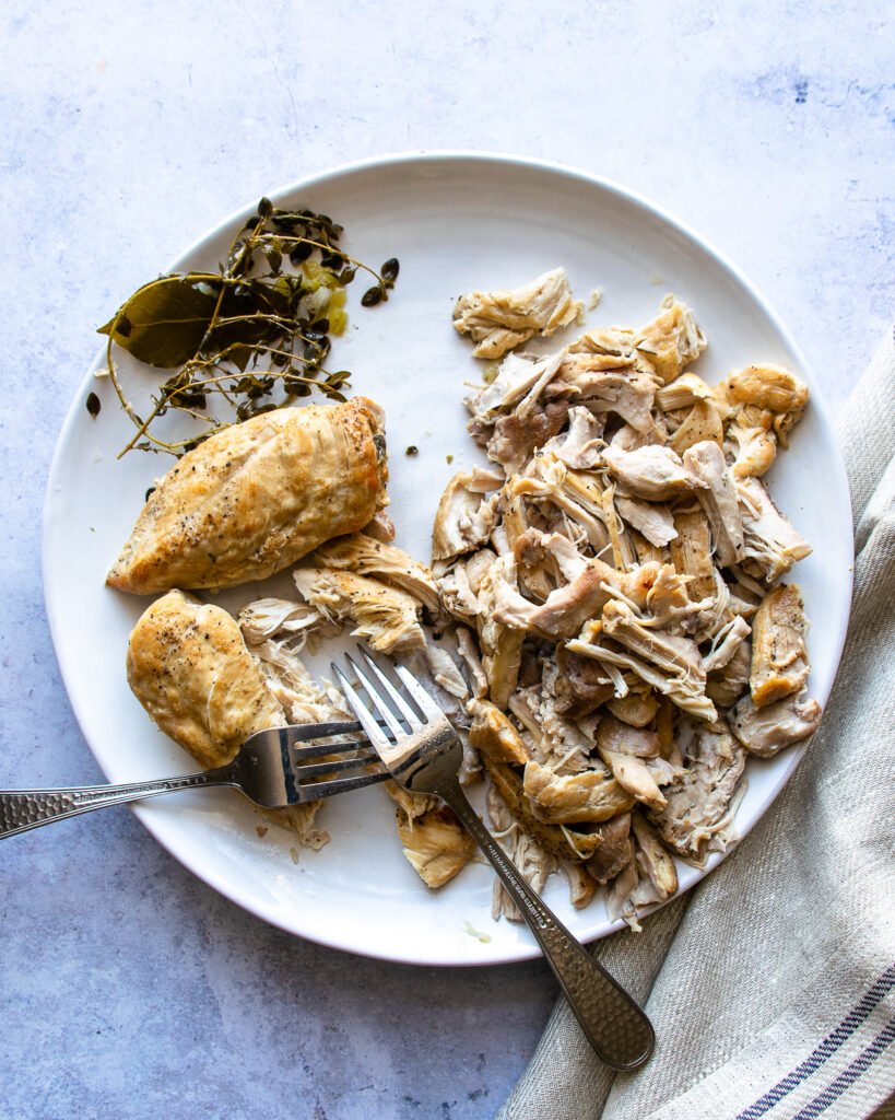 A white plate with cooked chicken being shredded by two forks.