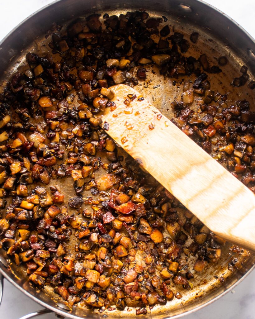 Close up of bacon, mushrooms and onions in a large saute pan with a wooden spoon.