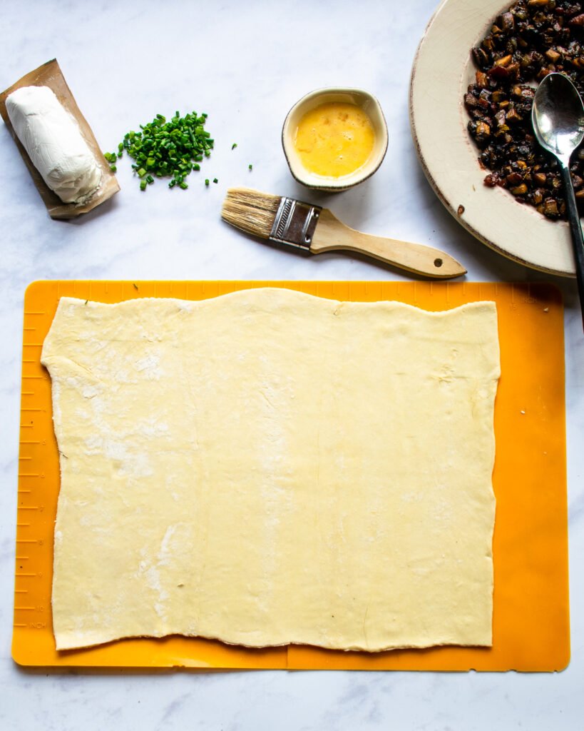A sheet of puff pastry on a silicone mat with goat cheese, egg wash, chives and a mushroom filling next to it.