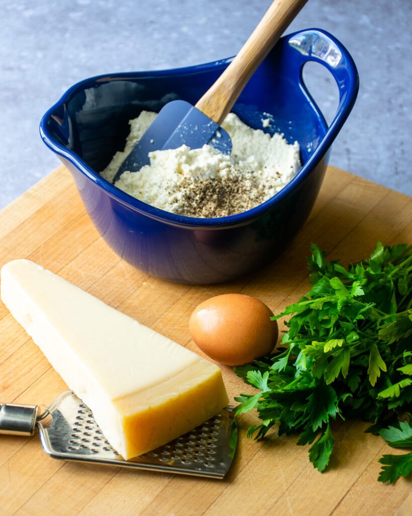 A blue bowl with ricotta cheese next to a piece of Parmesan cheese, an egg and some parsley.