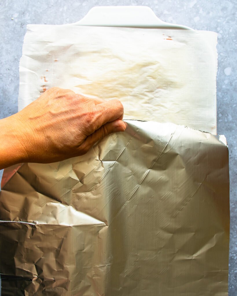 A hand covering a casserole dish with parchment paper and aluminum foil.