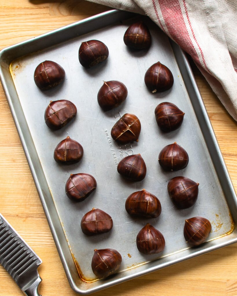 Scored chestnuts on a baking sheet with a knife and kitchen towel near by.