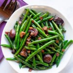 Green beans with onions and bacon in a white bowl with serving utensils and a purple napkin.