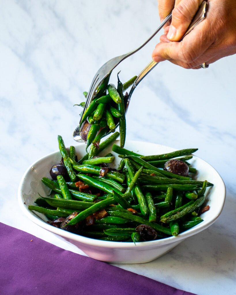 Hands serving green beans with onions and bacon out of a white bowl with serving utensils and a purple napkin.