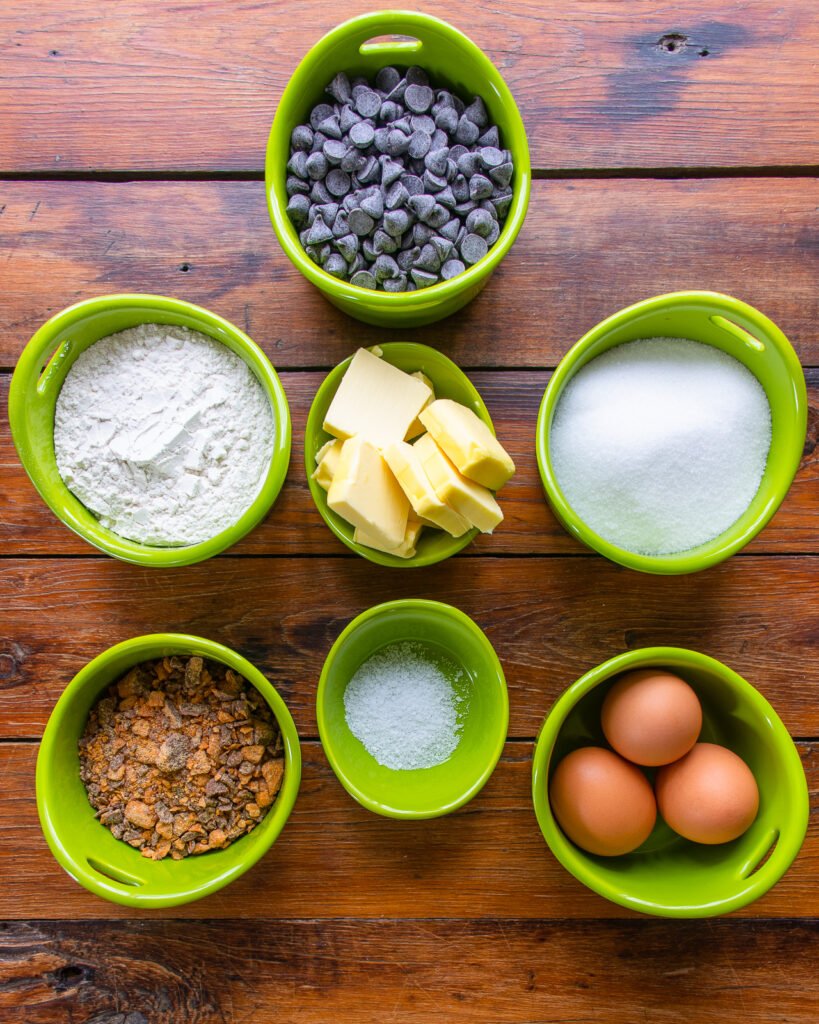 Ingredients in green ramekins on a wooden table - chocolate chips, flour, butter, sugar, caramel bits, salt and eggs.