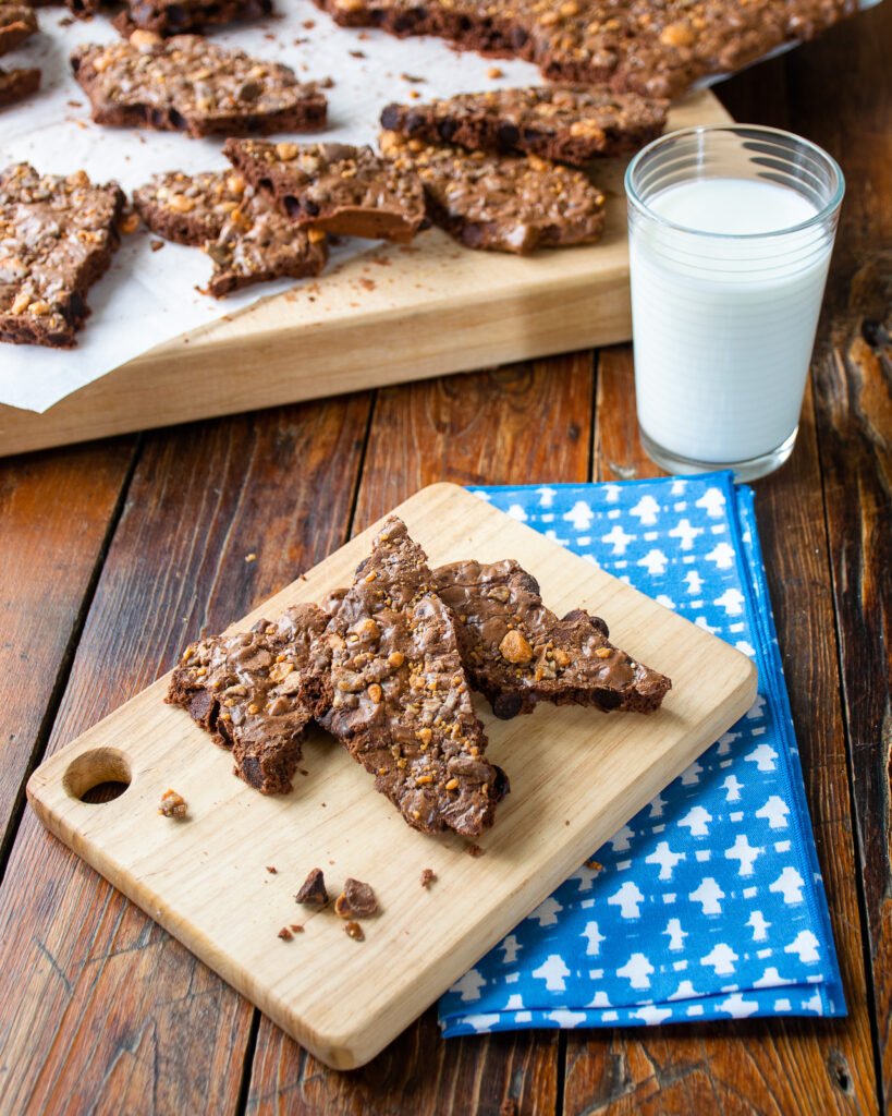 Three pieces of brownie brittle on a small wooden cutting board with a glass of milk and more brownie brittle in the background.