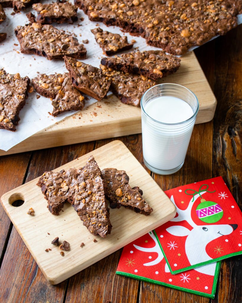 Three pieces of brownie brittle on a wooden cutting board with a glass of milk and a holiday napkin.
