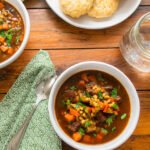 A white bowl of beef barley soup on a wooden table with a green napkin, spoon, glass, plate of biscuits and another bowl of soup.