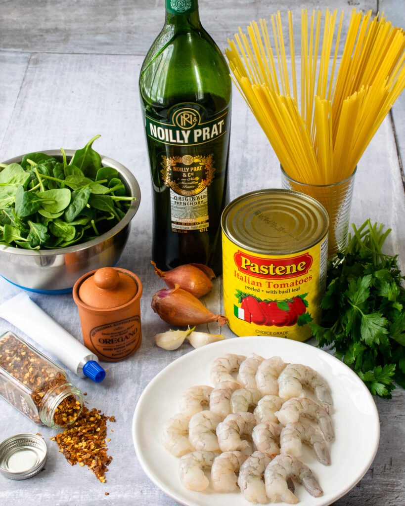 Ingredients on a table - shrimp on a plate, can of tomatoes, shallots, garlic, spices, spinach, dry vermouth and pasta.