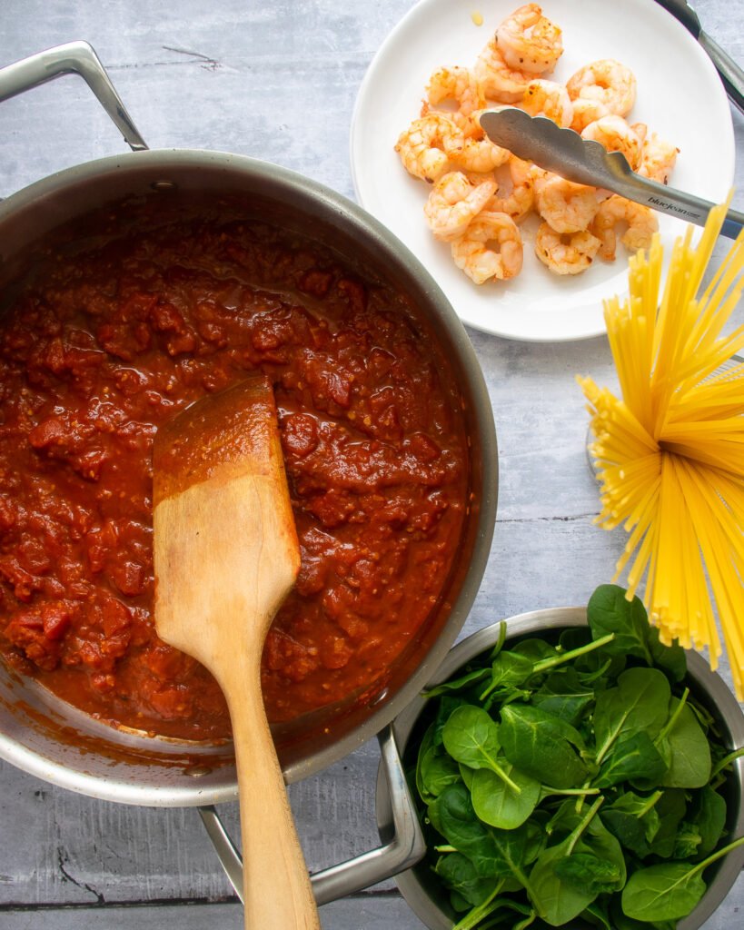 A saute pan with marinara with a bowl of spinach, a plate of seared shrimp and some linguine.