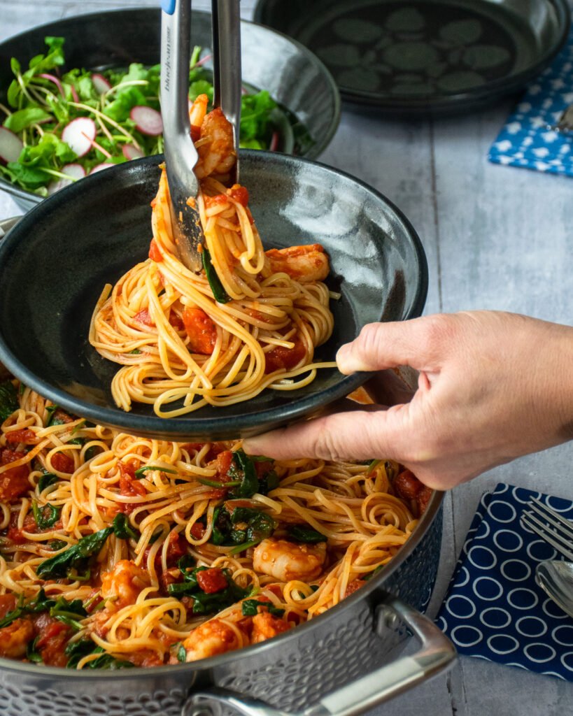 Hands serving up a portion of shrimp fra diavolo into a dark bowl.