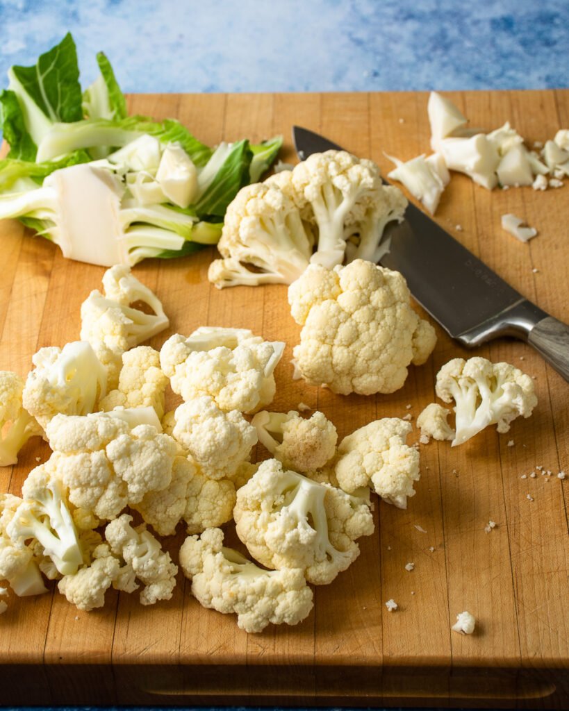 Cauliflower cut up on a wooden cutting board with a chef's knife.