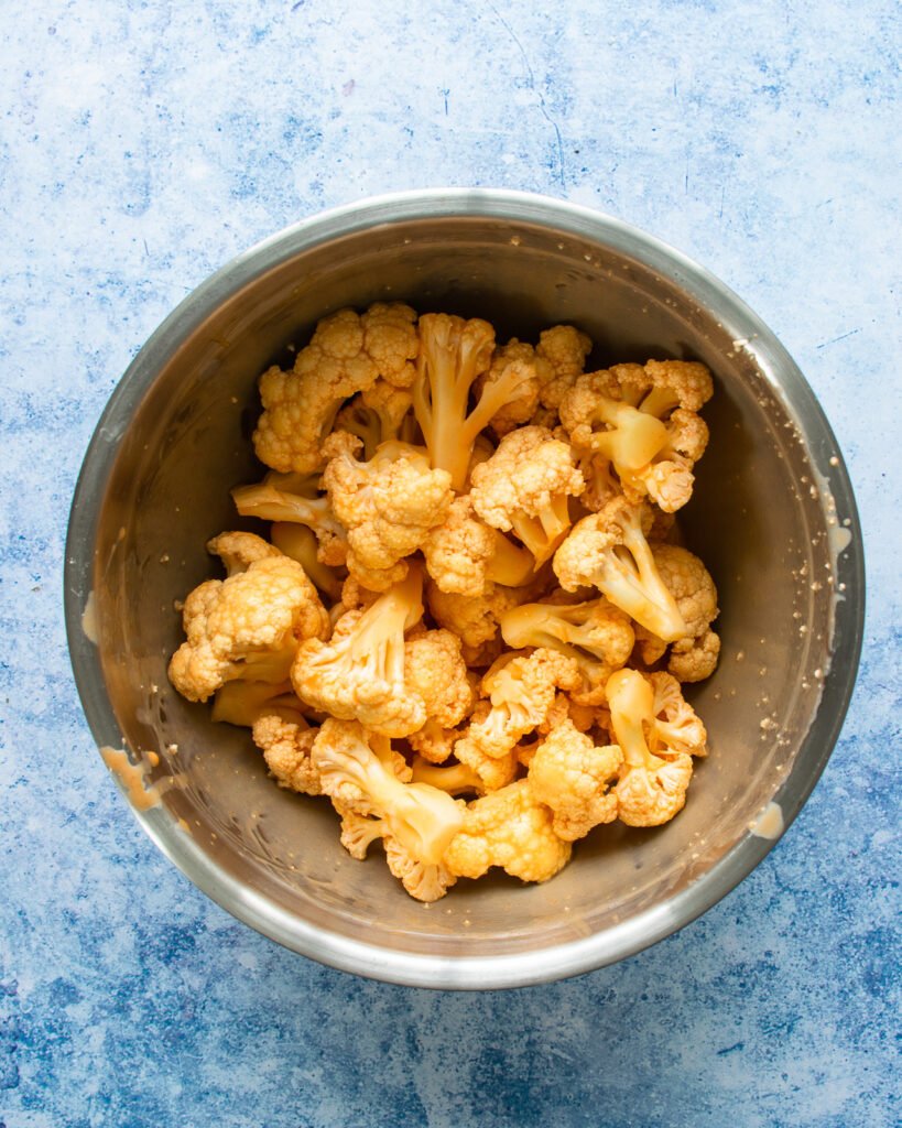 Cauliflower florets soaking in buttermilk and hot sauce in a stainless steel bowl with a blue background.