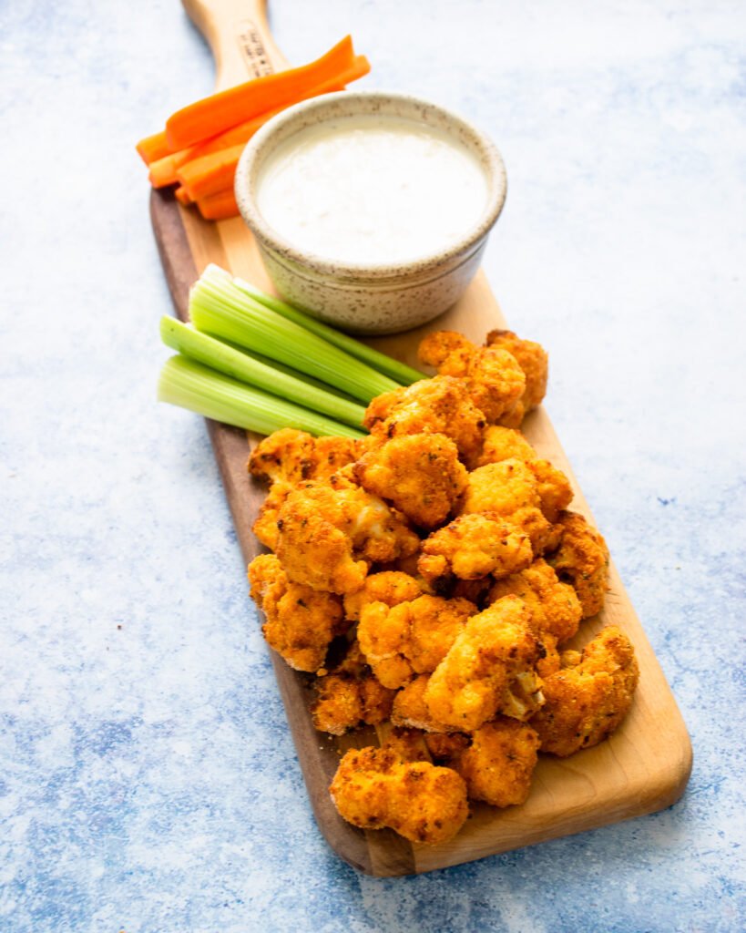Air fried Buffalo Cauliflower on a wooden board with carrot and celery sticks and blue cheese dressing with a blue background.