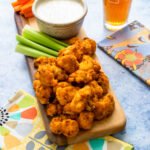 Air Fried Buffalo Cauliflower on a wooden board with carrot and celery sticks and blue cheese dressing, all on a blue countertop with a beer in the background.