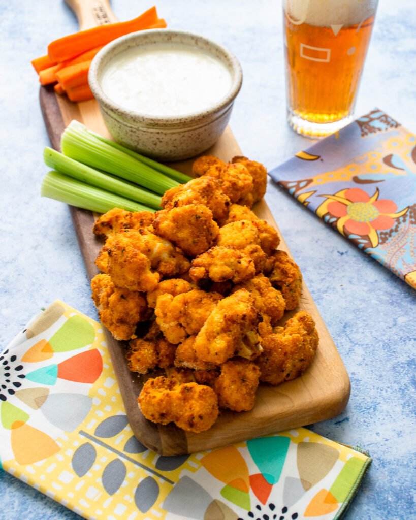 Air Fried Buffalo Cauliflower on a wooden board with carrot and celery sticks and blue cheese dressing, all on a blue countertop with a beer in the background.