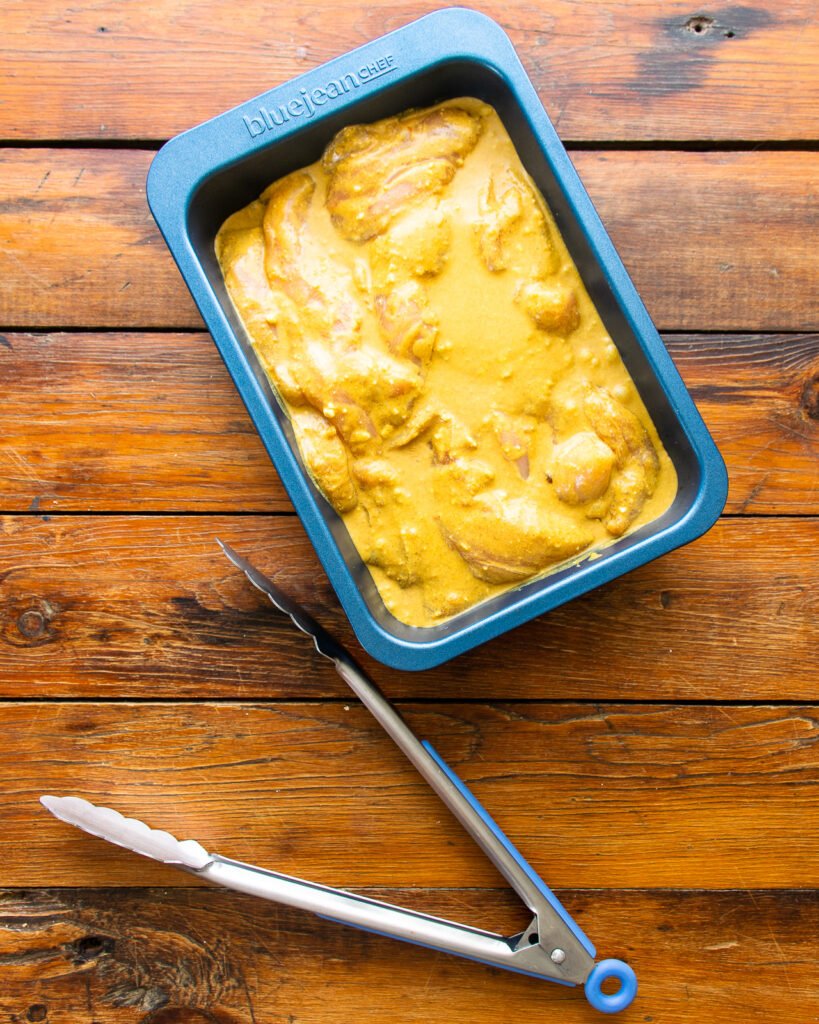 Chicken strips marinating in a blue baking pan on a wooden table with a pair of tongs next to it.