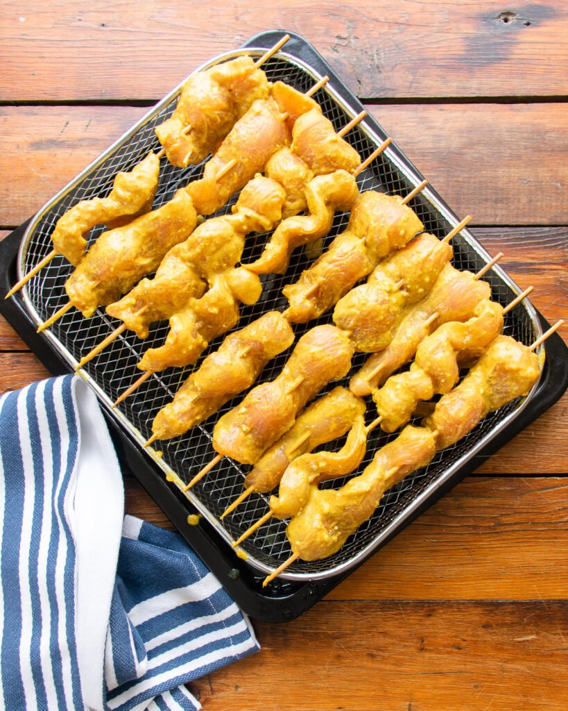 skewered chicken slices on an air fryer basket on a wooden table with a blue and white striped towel.