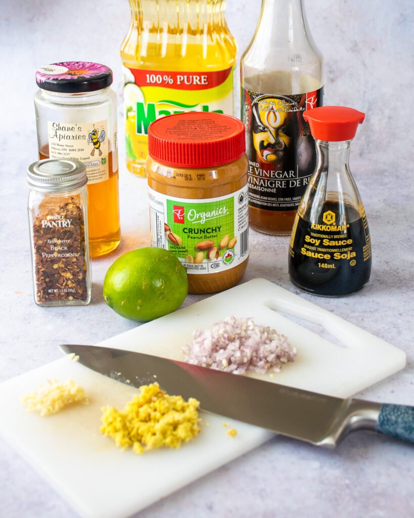 Ingredients on a counter with a cutting board and knife in front - peanut butter, soy sauce, chili flakes, honey, lime, rice vinegar and vegetable oil.