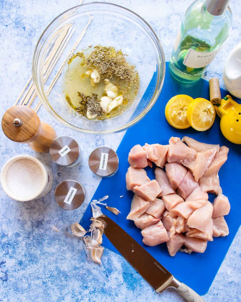 Ingredients on a blue cutting board on a blue countertop - chicken cubes, herbs, lemon, salt, pepper and olive oil.