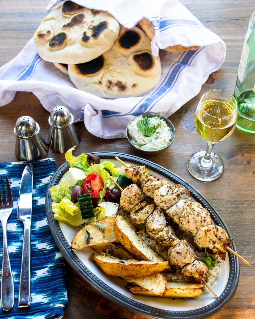 Chicken souvlaki on a plate with Greek potatoes and salad with a place setting on a wooden table.