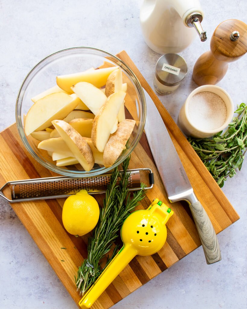 Ingredients on a striped wooden cutting board - potato wedges in a glass bowl, lemon, rosemary, oregano, salt, pepper, olive oil.