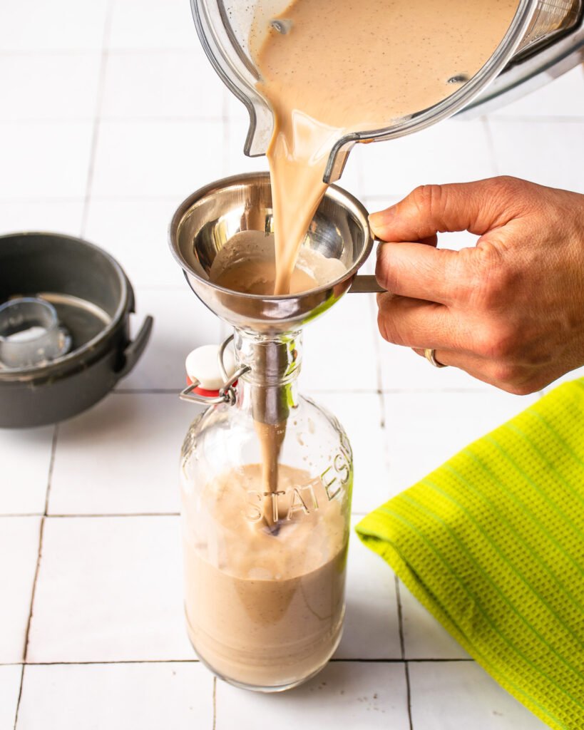 Pouring homemade Irish cream from a blender through a funnel and into a bottle on a white tiled counter with a green towel nearby.