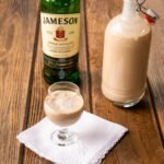 A bottle of Jameson's Irish Whiskey next to a bottle of homemade Irish cream and a small glass of Irish cream on a wooden table.
