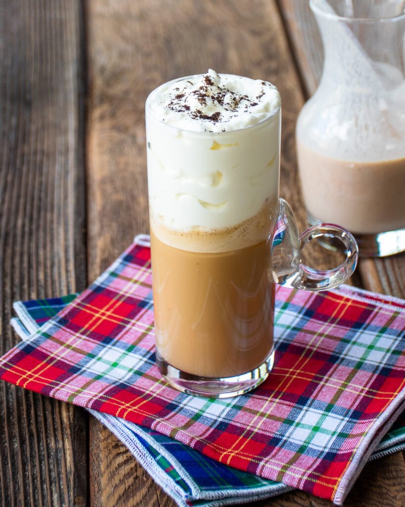 An Irish coffee in a glass resting on a plaid napkin on a wooden table with a small carafe of Irish cream in the background.