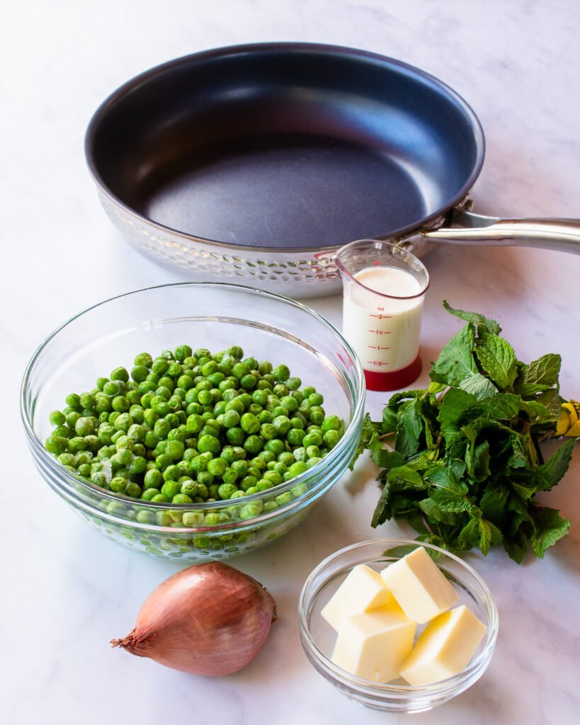 Ingredients on a marble countertop - a skillet, peas, shallot, heavy cream, butter and mint.